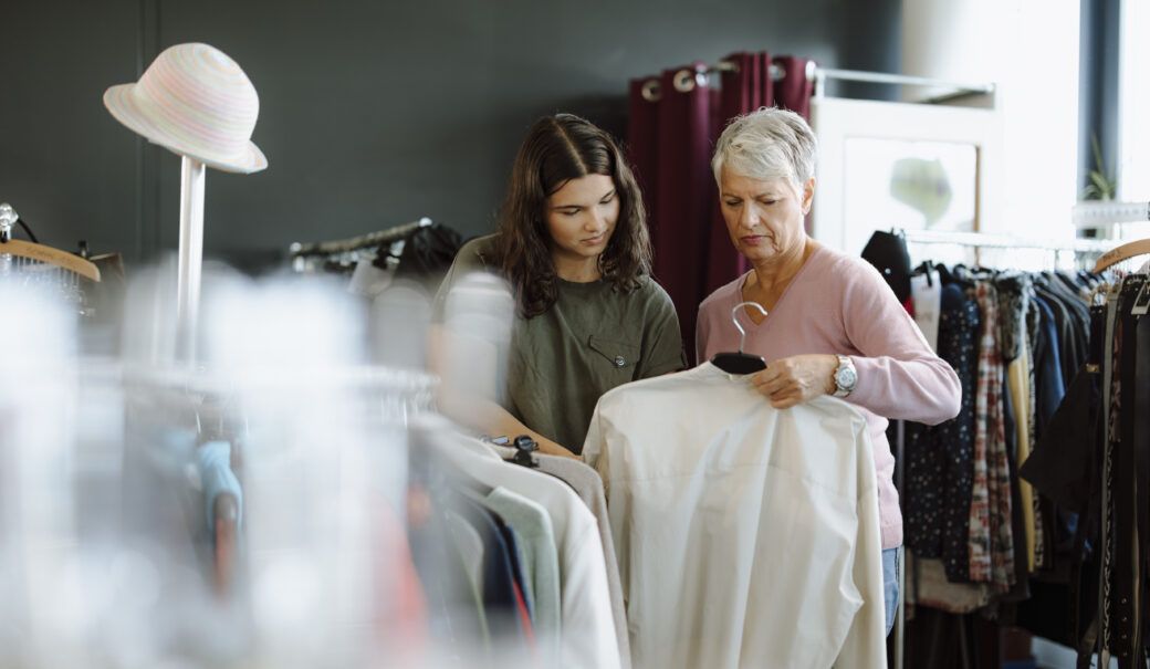 Eine &auml;ltere Dame mit grauem Haar steht in einem Secondhandladen und h&auml;lt eine wei&szlig;e Bluse in der Hand. Neben ihr steht ein junges M&auml;dchen mit lockigem Haar und einem olivgr&uuml;nen T-Shirt, das ebenfalls die Bluse interessiert betrachtet.