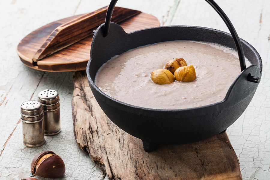 Schwarzer Eisenkessel mit Henkel gefüllt mit cremiger Maronisuppe. In der Suppe sind drei ganze Maroni zu sehen. Der Kessel steht auf einem Holzbrett, daneben silberne Salz- und Pfefferstreuer