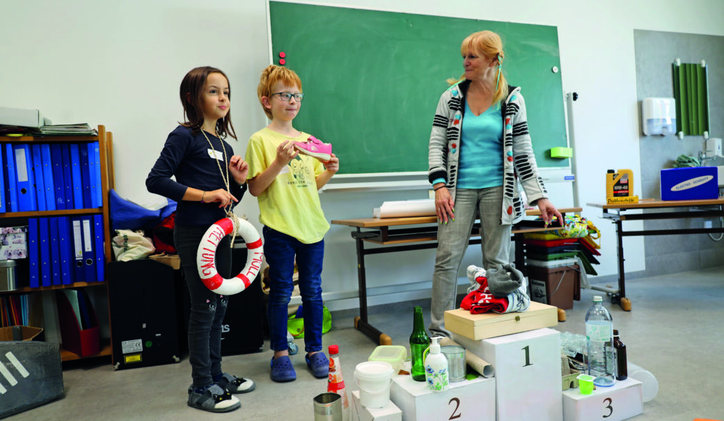 Zwei Kinder im Volksschulalter stehen neben einer bloden Frau mit geflochtenen Zöpfen und einer Schleife im Haar. Davor ein Siegespodest mit Abfällen darauf und im Hintergrund eine Tafel und ein Regal mit blauen Ordnern.