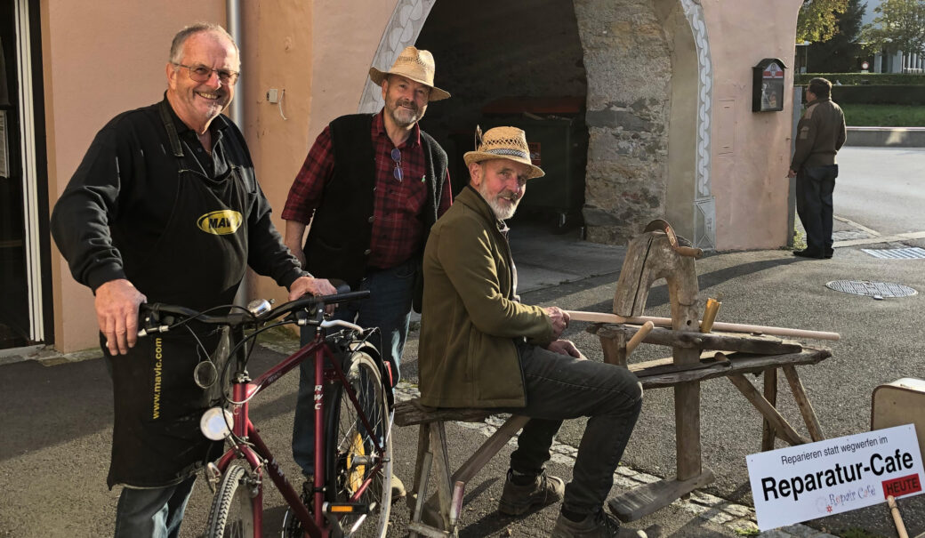 Auf den Foto sind drei Herren im mittleren mit höheren Alter zu sehen. Ein Herr auf der linken Seite hält eine Fahrrad. Ein zweiter Herr sitzt auf einem Holzblock und macht Holzarbeiten. Im rechten unterem Eck sieht man ein Schild auf dem Folgendes steht: Reparieren statt wegwerfen im Reparatur-Cafe.