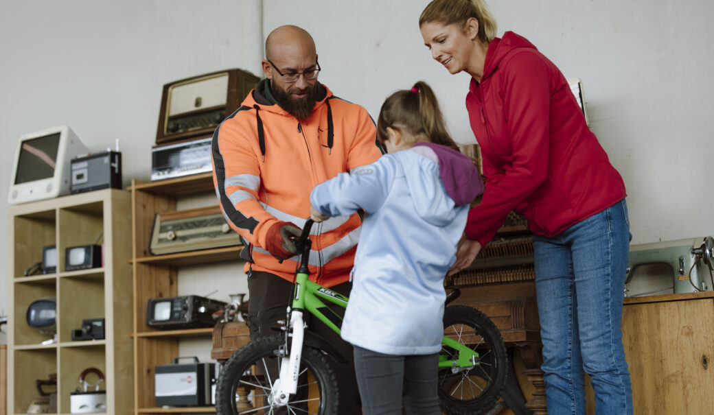 Ein kleines Mädchen bekommt in einem ReUse and Repair Shop ein abgegebenes Fahrrad von dem Mitarbeiter in die Hand gedrückt.